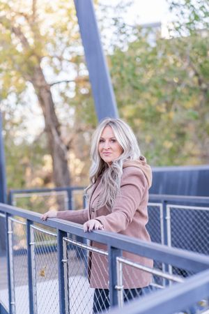Person in tan jacket leaning on metal railing of outdoor walkway during autumn season.