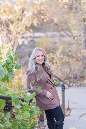 Person in brown coat poses outdoors against autumn foliage and green plants.