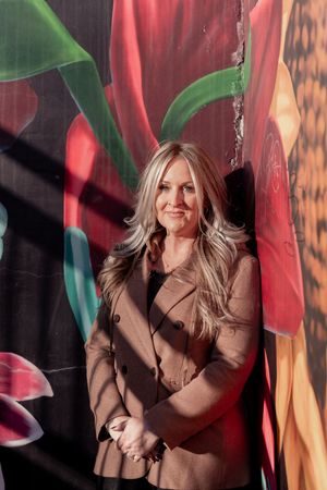 A person in a brown blazer poses against a backdrop of red and green abstract light patterns.