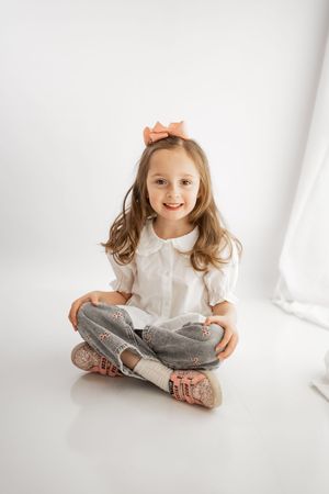 Smiling young girl with pink bow sitting cross-legged on white floor in white blouse and gray jeans.