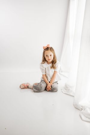 Young girl with pink bow sits cross-legged on white floor wearing white top and gray jeans in bright studio setting.