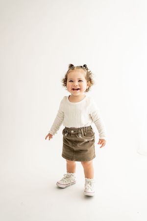 Small child wearing white long sleeve shirt, brown skirt and white sneakers stands smiling against white background.
