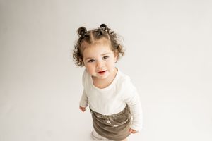 A small child in a white long sleeve shirt and gray skirt looks up with curly hair in buns against a light background.