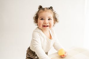 A joyful toddler in a white sweater holds a yellow cup while sitting against a white background.