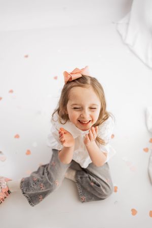 A joyful little girl with a pink bow sits on a white floor surrounded by heart confetti, clapping and laughing happily.