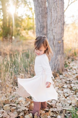 Young girl in white dress twirling among autumn leaves in sunlit forest.