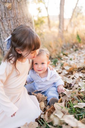 A young girl kisses a baby sibling sitting in autumn leaves beside a tree in golden sunlight.