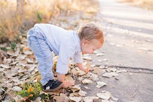 Toddler in jeans and white shirt bending down to pick up fallen autumn leaves on a sunlit path.