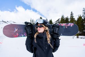 Snowboarder in black winter gear holds up two snowboards against snowy mountain backdrop and blue sky.