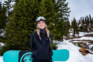 A snowboarder rides down a snowy mountain slope surrounded by evergreen trees on a bright winter day.