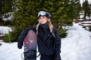 Skier in black winter gear smiles while posing on a snowy slope with evergreen trees in the background.