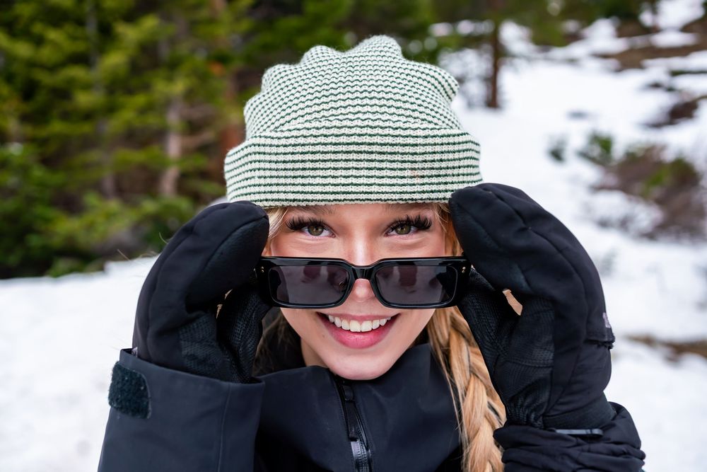 Winter portrait of someone wearing a striped beanie hat, sunglasses and black winter coat in snowy outdoor setting.