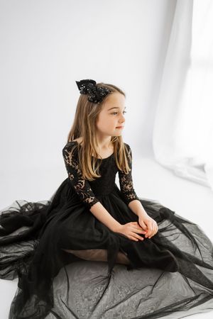 Young girl in elegant black lace tulle dress with bow headband sits gracefully on white studio floor.