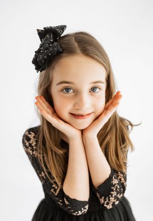 Young girl in black lace dress with bow headband, smiling with hands on cheeks against white background.
