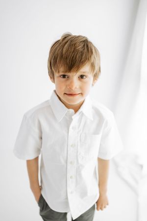 A young boy with blonde hair wearing a white button-up short sleeve shirt smiles softly against a white background.