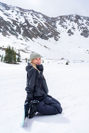 A person in winter gear poses playfully in the snow with snow-capped mountains and evergreen trees in the background.