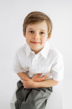 A young boy in a white shirt and olive pants sits smiling with hands clasped against a white background.