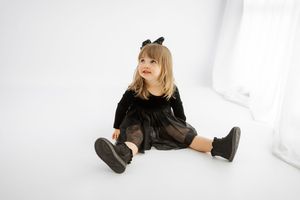 Young girl in black velvet dress and boots with bow headband sitting on white floor.