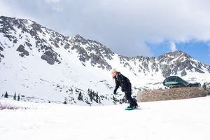Skiers navigate through snowy mountain terrain with snow-capped peaks and blue sky in the background.