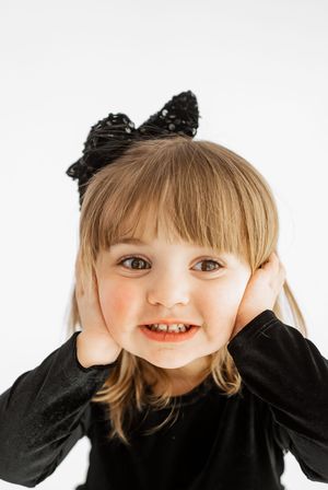 Cute toddler girl in black outfit with cat ear headband smiling at camera against white background.