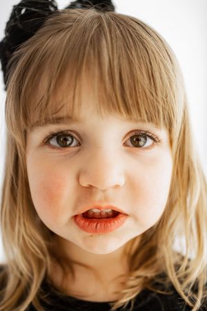 Close-up portrait of a young blonde girl with brown eyes wearing a black bow, looking at the camera with a curious expression.