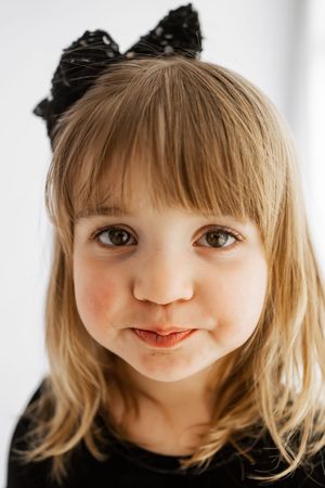 A young girl with blonde hair and a black bow headband wearing a black top, smiling softly at the camera.