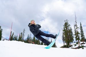 A snowboarder soars through the air against a backdrop of evergreen trees and cloudy sky at a winter resort.