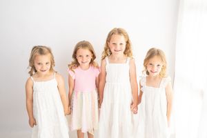 Four young girls in white and pink dresses hold hands and smile together in a bright white studio setting.