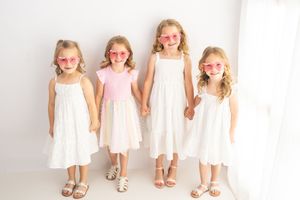 Four young girls in white dresses and pink heart sunglasses holding hands and smiling in a bright white studio.