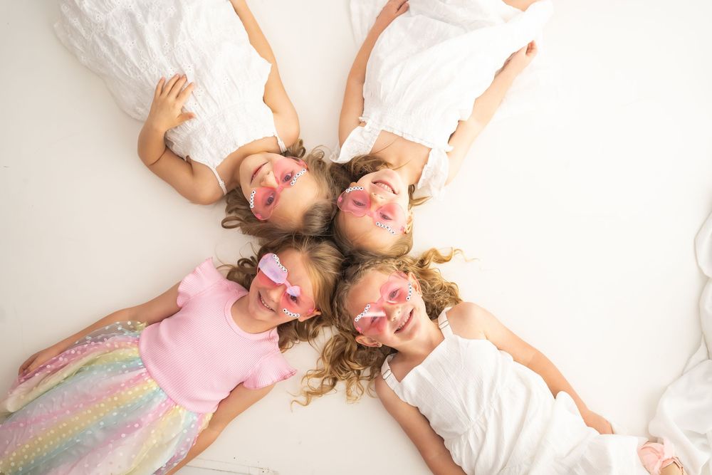 Four young girls lying in a circle with heads together, wearing pink butterfly eye masks at a fun sleepover party.