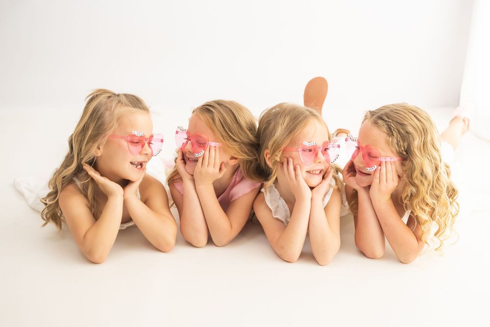 Four young girls in pink heart sunglasses lying on white floor, smiling and posing with hands on cheeks.