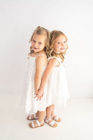 Two young girls in white dresses and sandals standing back-to-back, smiling and holding hands against a white background.