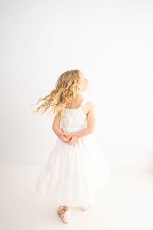 A young girl with flowing blonde curly hair twirls in a white dress and pink shoes against a white background.