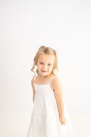 A smiling young girl with blonde curly pigtails wearing a white sundress poses against a white background.