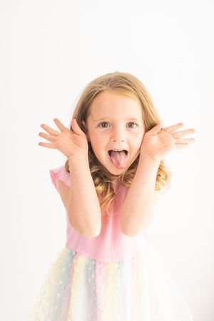 A playful young girl in a pink rainbow dress sticks out her tongue and fans her hands beside her face against a white background.
