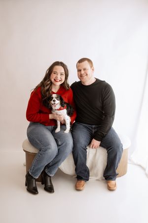 A couple sits on a white backdrop with their small black and white dog wearing a red sweater in a photo series.