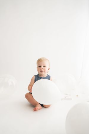 Baby in denim romper plays with large white balloon on light background in minimalist photo series.