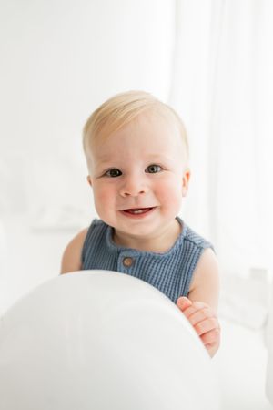 Happy baby in blue sleeveless romper smiling while holding white balloon against light background.