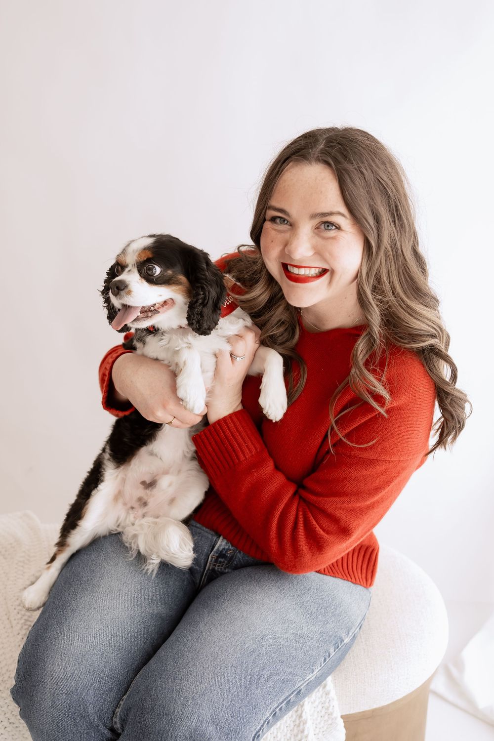 Person in red sweater and jeans playing with small white and brown dog in bright indoor setting.