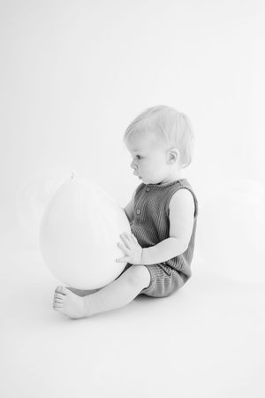 Black and white portrait of a baby in striped overalls sitting on white background with bare feet.