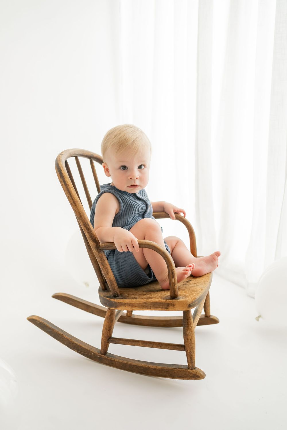 Small child sits in a vintage wooden rocking chair against a white curtain backdrop.