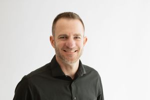 Professional headshot of someone wearing a dark jacket against a white background with a warm, friendly expression.