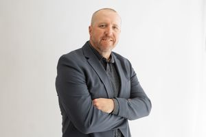 Professional portrait of someone in a dark blazer and shirt standing with arms crossed against a white background.