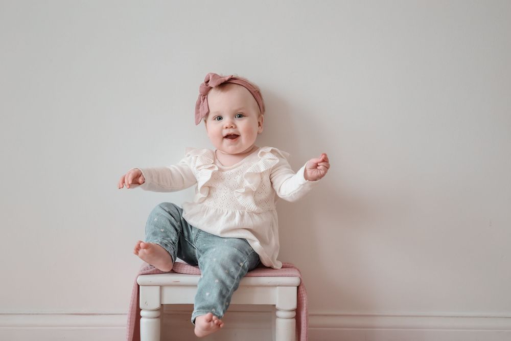 Baby wearing pink bow and ruffled white top sits on small white chair.