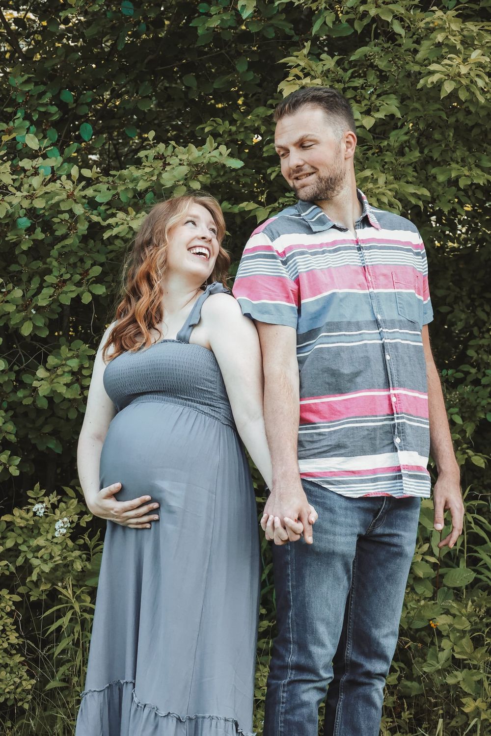 Expecting couple sharing laugh together against green foliage background.