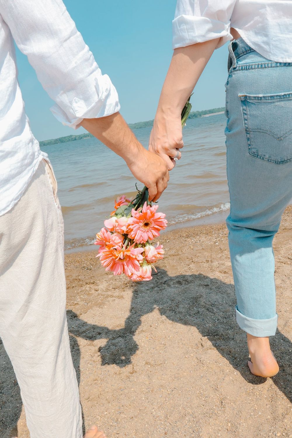 Two people holding hands on beach with coral flowers against turquoise ocean background.