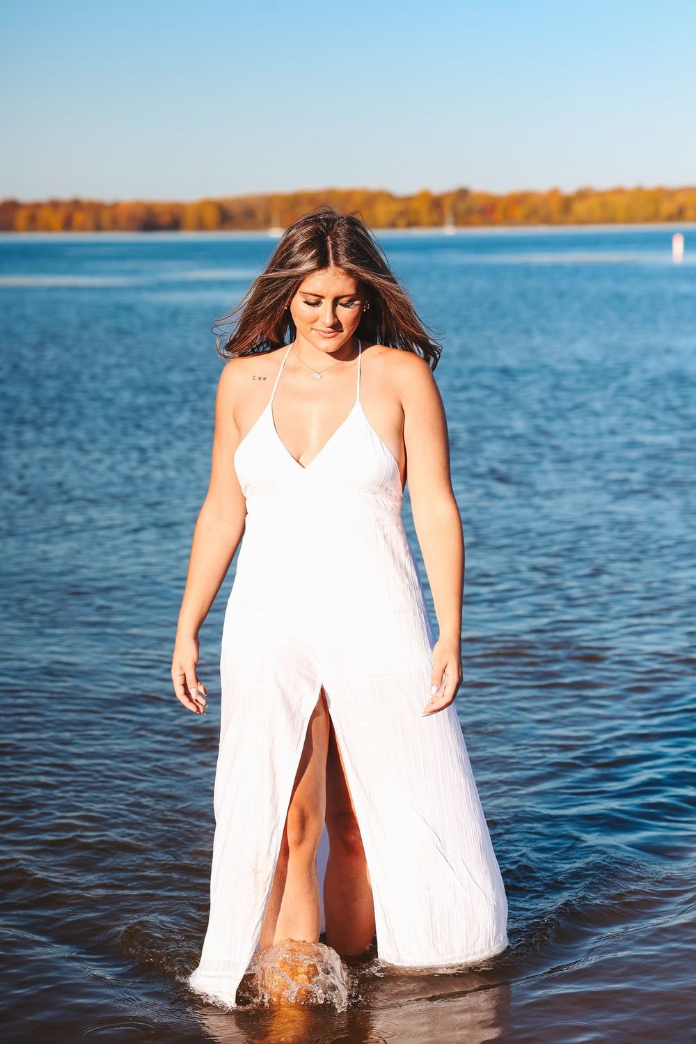 Model in white sundress splashing in lake water during sunset photoshoot.