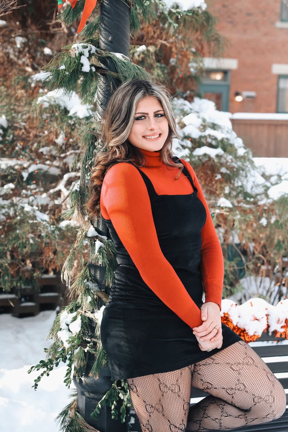 Winter portrait of person in orange turtleneck and black dress against snowy evergreen backdrop.