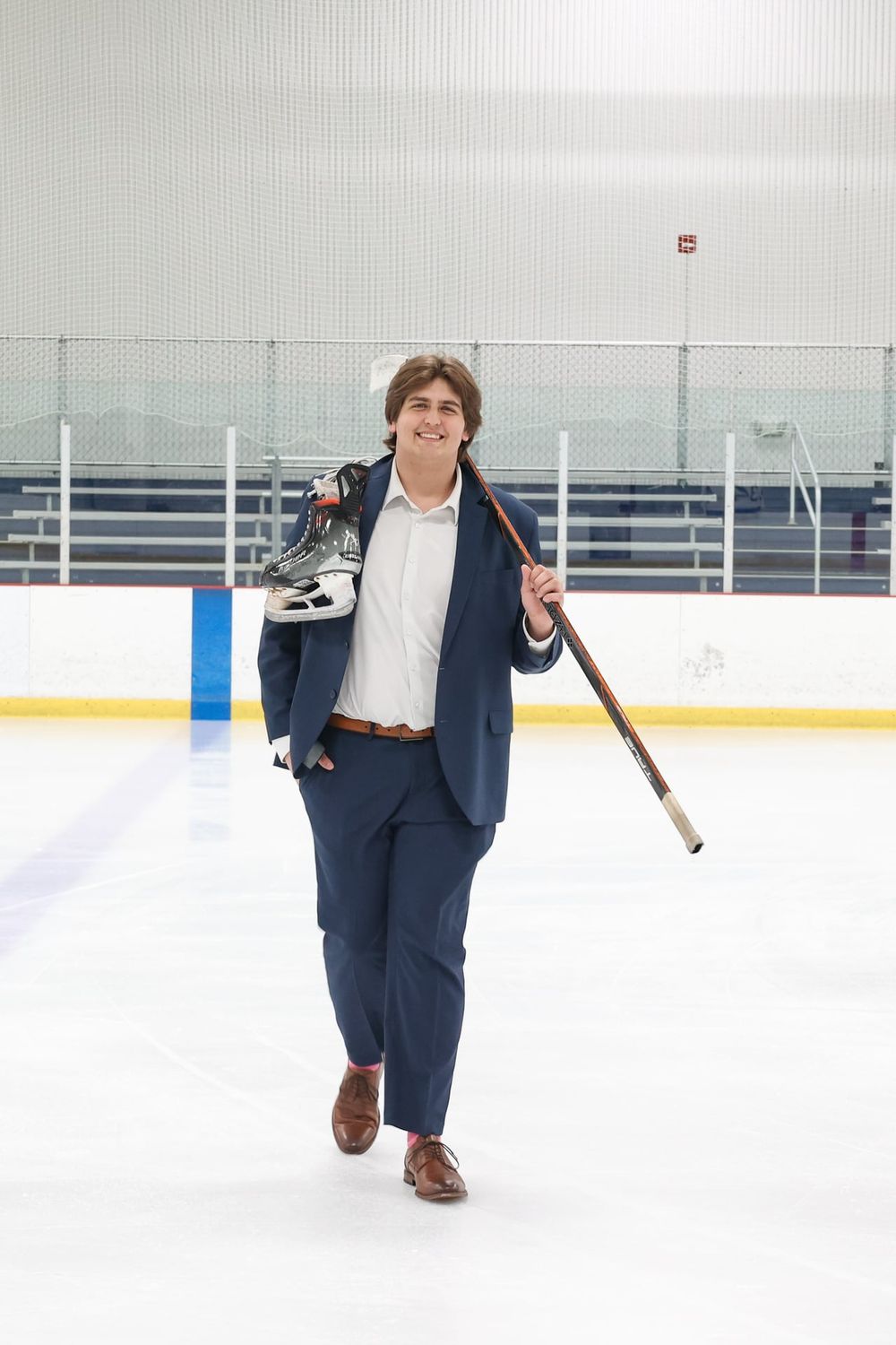 Person in business suit carrying hockey equipment while walking on ice rink.
