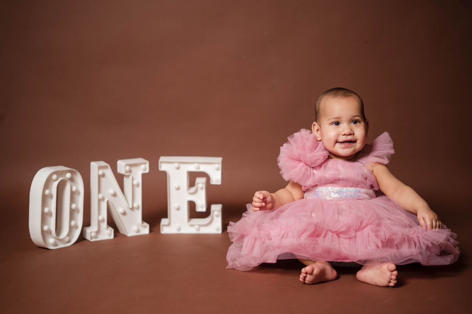 Adorable baby girl in pink ruffle dress sitting beside illuminated ONE marquee letters for first birthday.